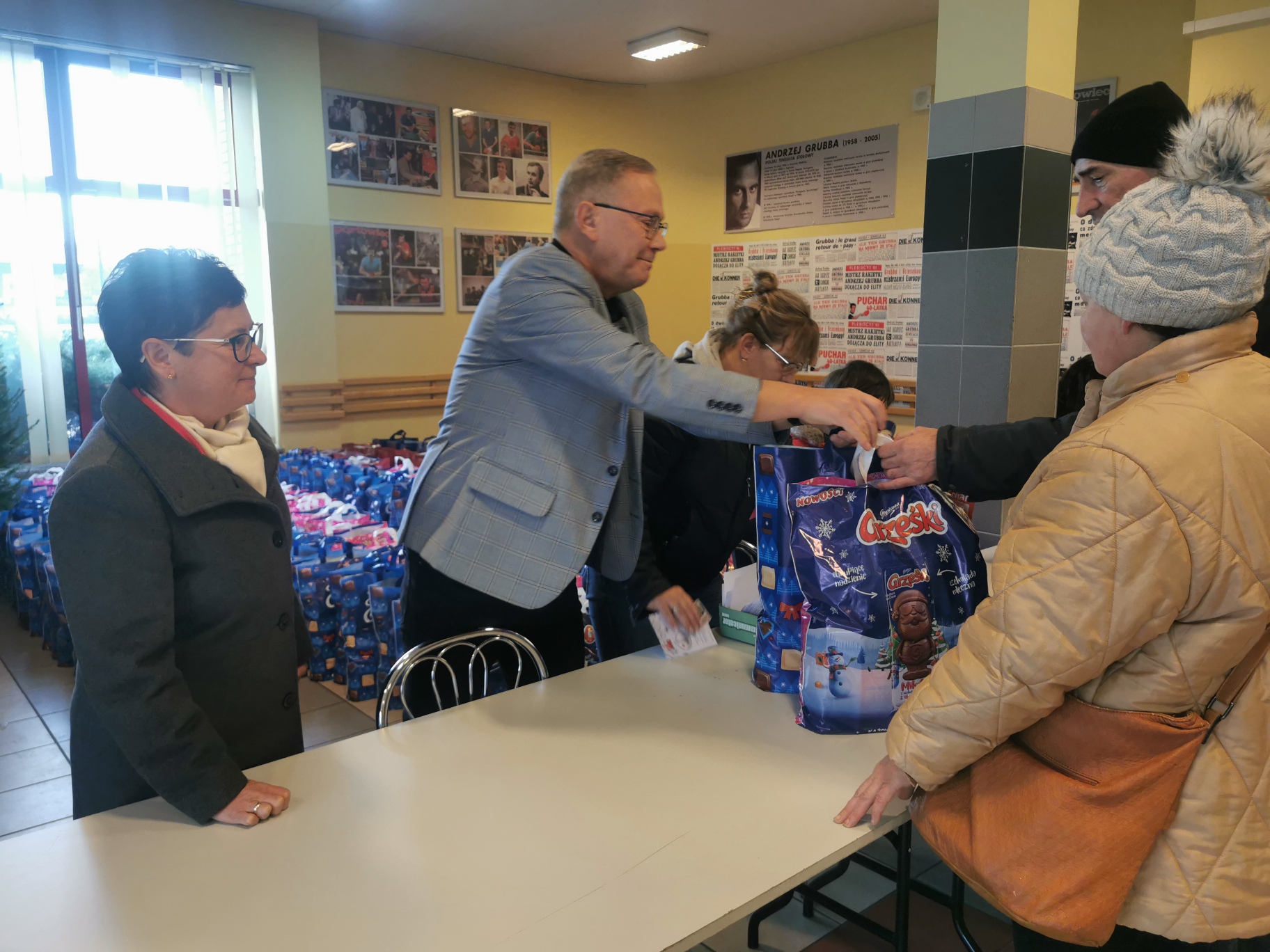 Dyrektor Ośrodka i Prezydentem miasta stoja za stołem. Prezydent pochylając się przekazuje paczkę mężczyźnie.