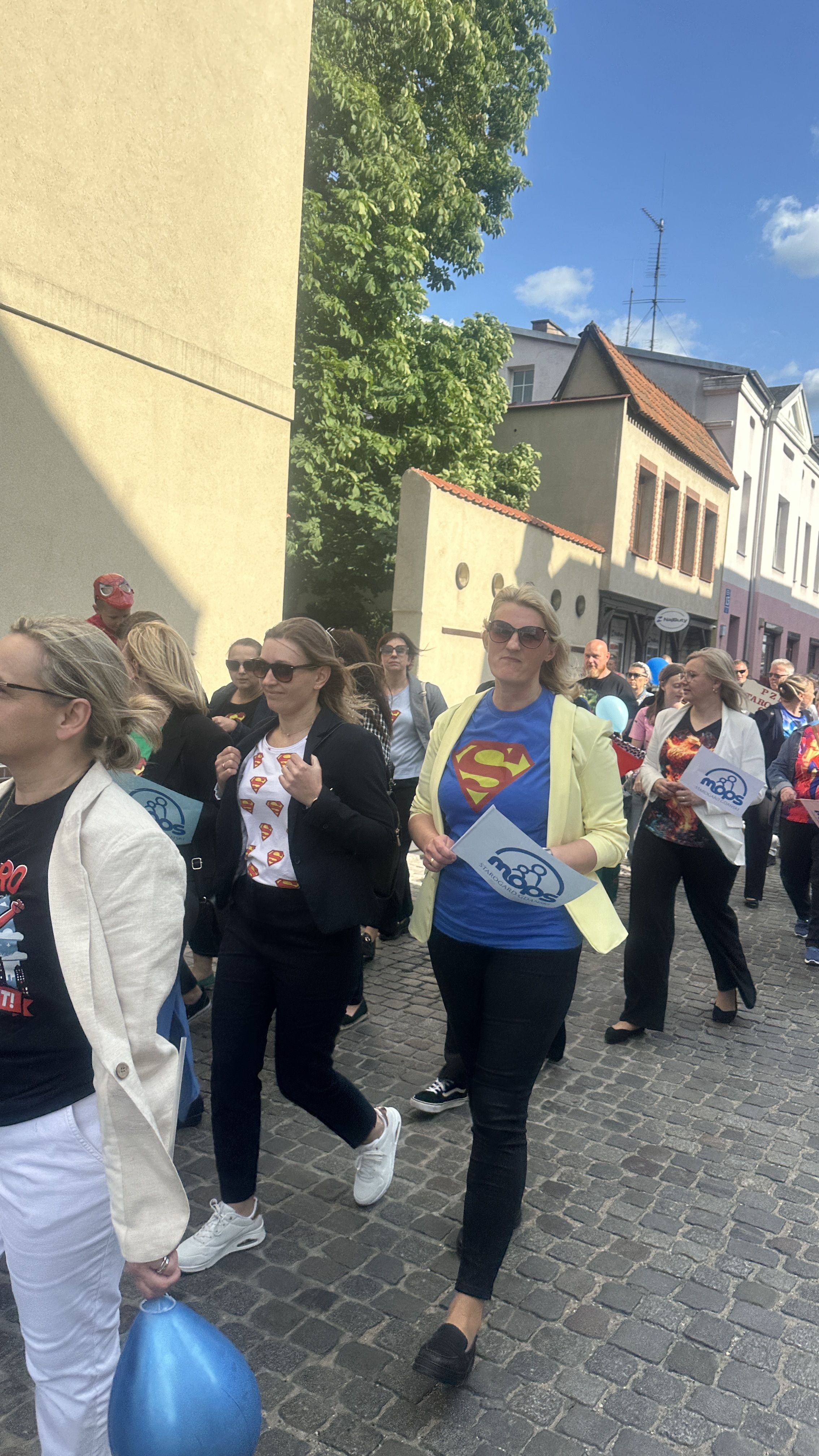 Grupa osób idąca po kostce brukowej trzymająca w rękach balony lub male flagi z logiem oraz napisem MOPS.